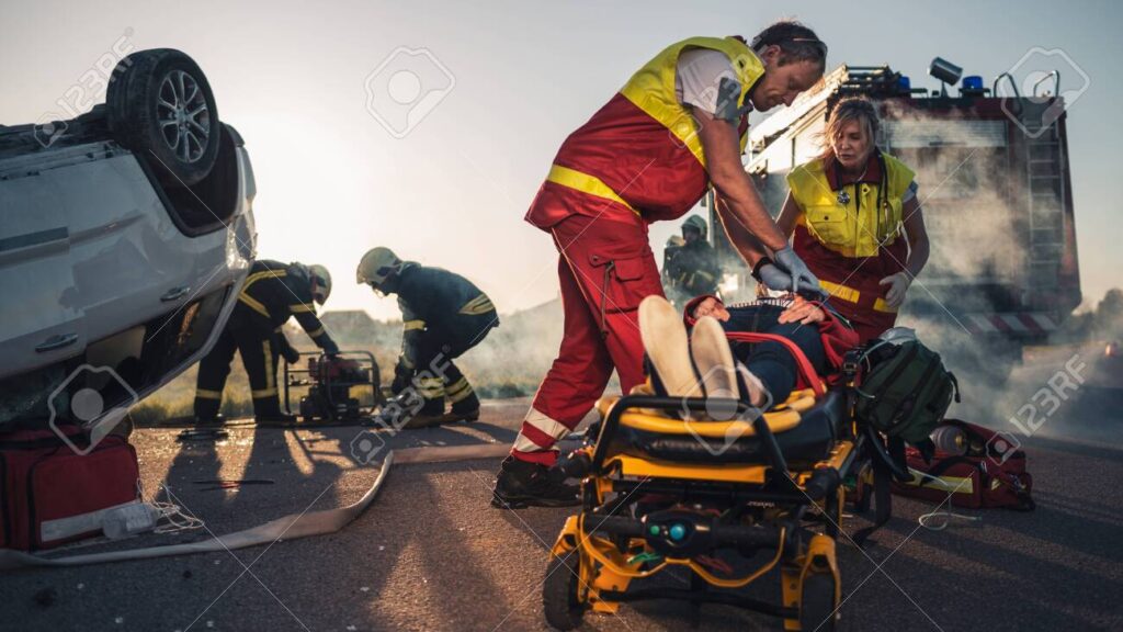On the Car Crash Traffic Accident Scene: Paramedics Saving Life of a Female Victim who is Lying on Stretchers. They Apply Oxygen Mask, Do Cardiopulmonary Resuscitation CPR and Perform First Aid